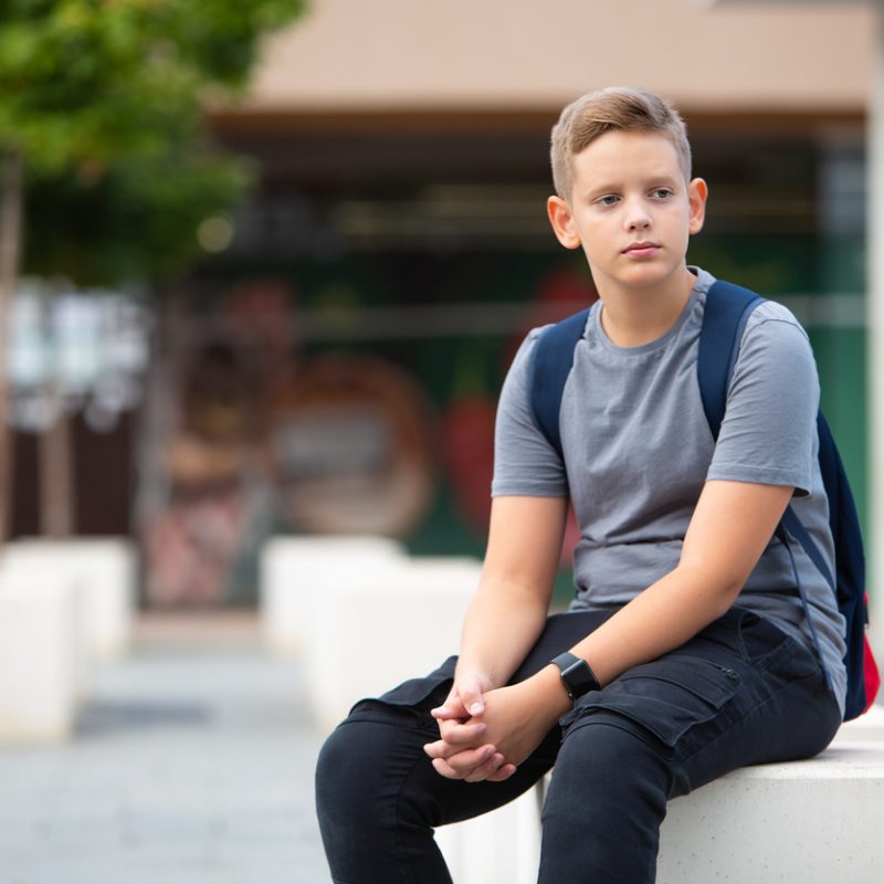boy sitting outside school with somber expression, wondering how does school affect teenage mental health