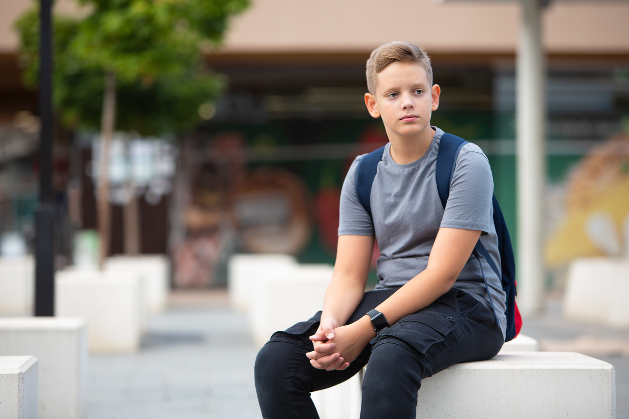 boy sitting outside school with somber expression, wondering how does school affect teenage mental health